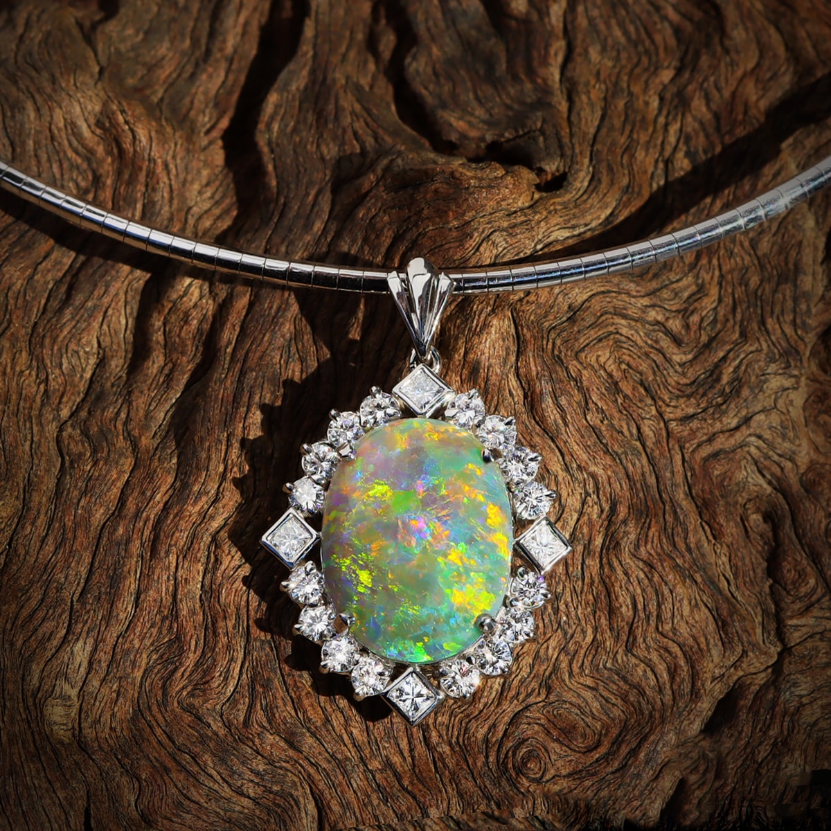 A pendant consisting of an oval-shaped solid crystal opal, surrounded by a halo of white diamonds, set in a platinum setting. The Australian opal is a high dome stone, with a flagstone pattern and vibrant flashes of greens, oranges, and yellows. The pendant includes a white gold chain and is resting on a dark wood background.