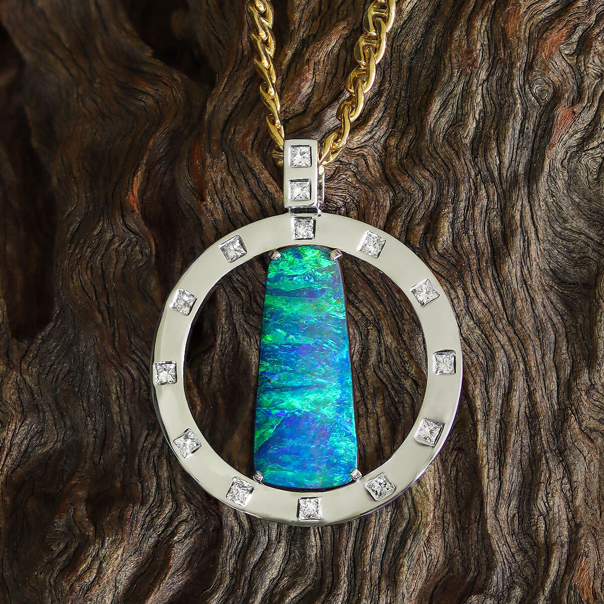 The round pendant consists of a flat platinum ring, and a rectangular boulder opal vertically set in the ring by four claws. The opal is a gem-grade quality natural solid Australian boulder opal, with beautiful vivid blue and green horizontally striated pattern. The ring that encloses the opal is silver in colour, made of platinum and has 12 colourless high jewellery grade princess cut diamonds set all around it, just like the numbers on a clock. The bail is rectangular in shape and there’s two more princess cut diamonds set in it, one above another. Through the bail passes a thick 18K solid gold chain. This is a front on view of a boulder opal pendant resting on textured natural wood.