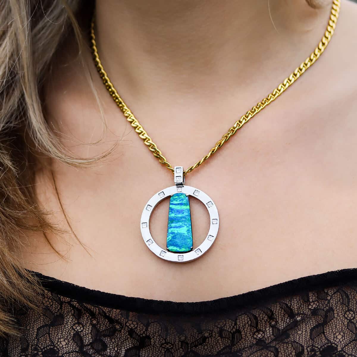 A close-up of a hand-crafted boulder opal pendant on a thick 18K solid gold chain around a young woman’s neck. The round pendant is photographed front on and the vivid blue and green tones of the Australian boulder opal contrast nicely with the platinum ring the rectangular opal is set vertically within.